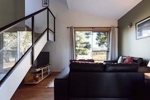 A living room with a black leather sofa, a television on a stand, a staircase, and a large window with curtains.