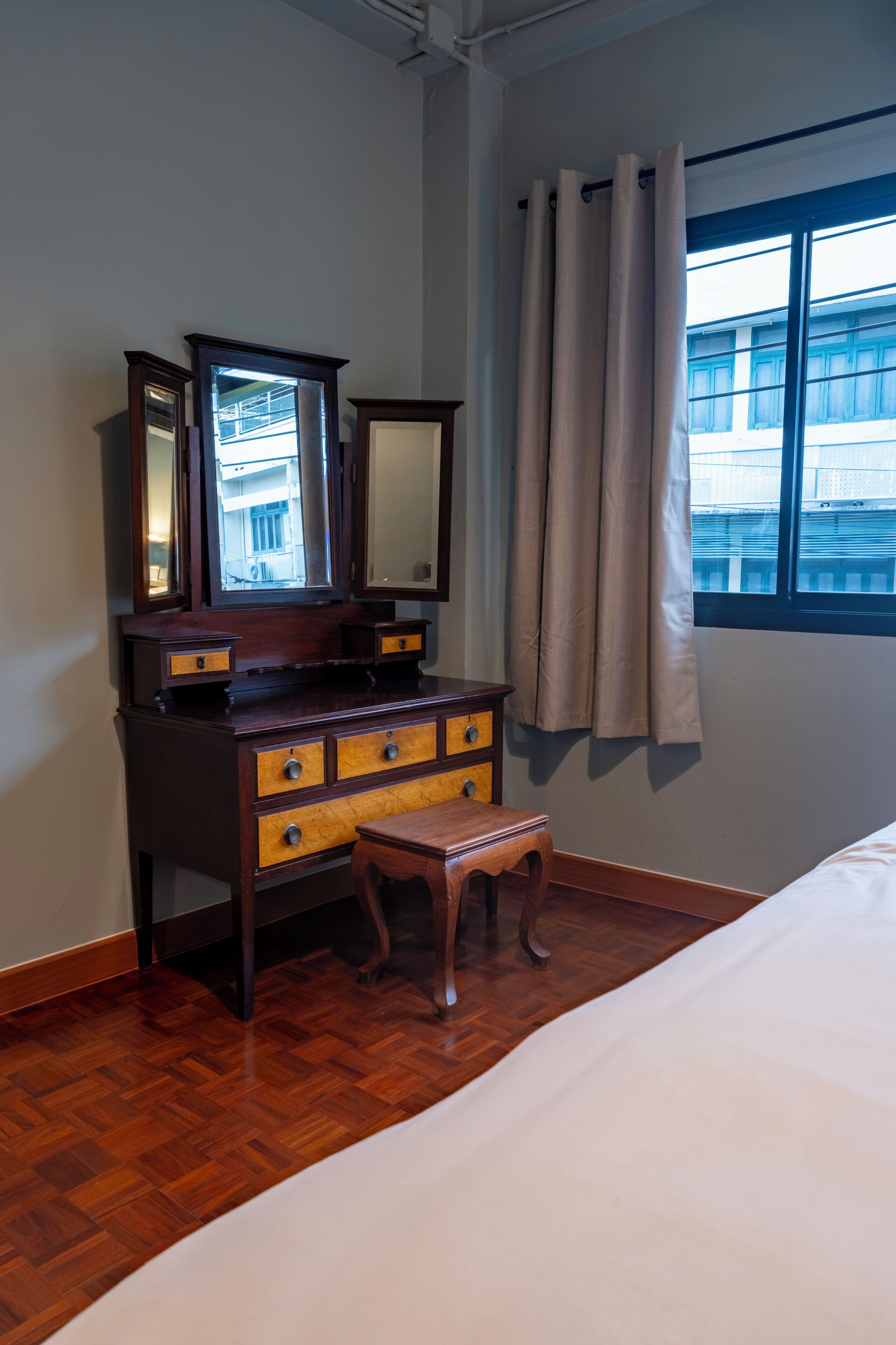 Ein Zimmer mit einem Holzkoffer, einem Spiegel, einem Fenster mit Vorhängen und einem Bett.