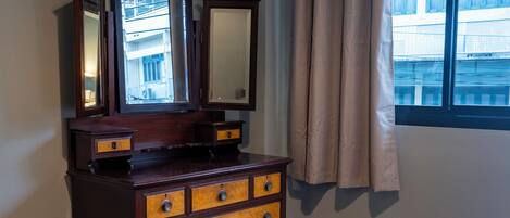 Ein Zimmer mit einem Holzkoffer, einem Spiegel, einem Fenster mit Vorhängen und einem Bett.
