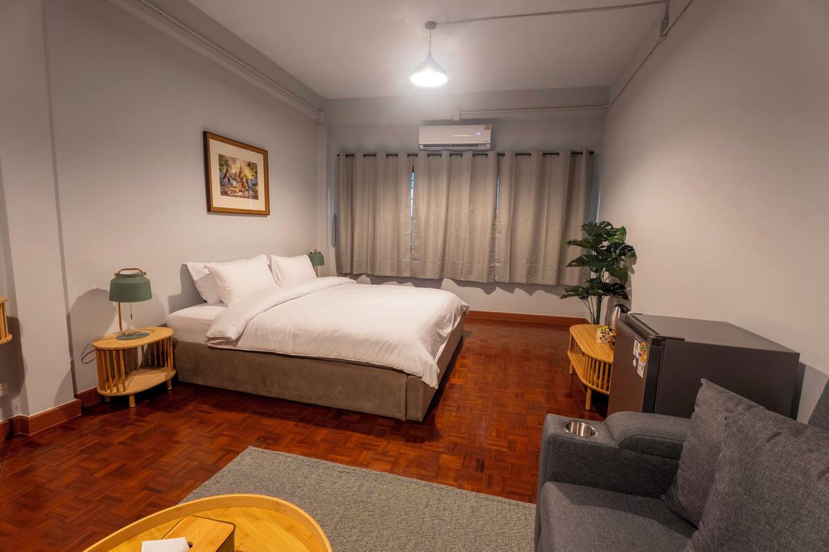 Une chambre à coucher avec un lit, un canapé gris, une table de chevet en bois, une lampe verte, un tableau au mur et un appareil de climatisation.