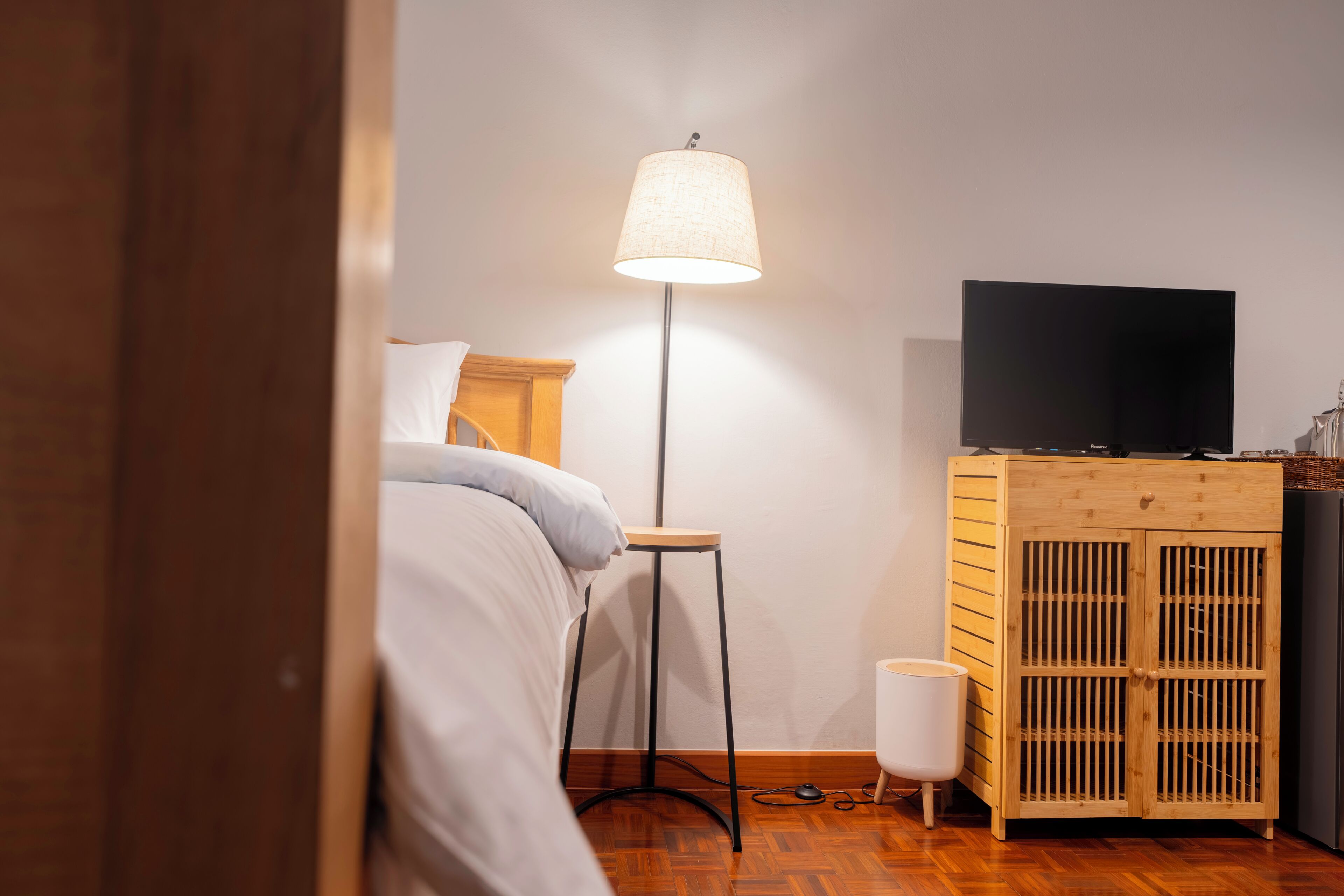 A bedroom with a bed, a standing lamp, a TV on a wooden cabinet, and a white trash bin.