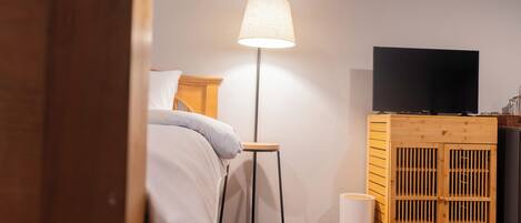 A bedroom with a bed, a standing lamp, a TV on a wooden cabinet, and a white trash bin.