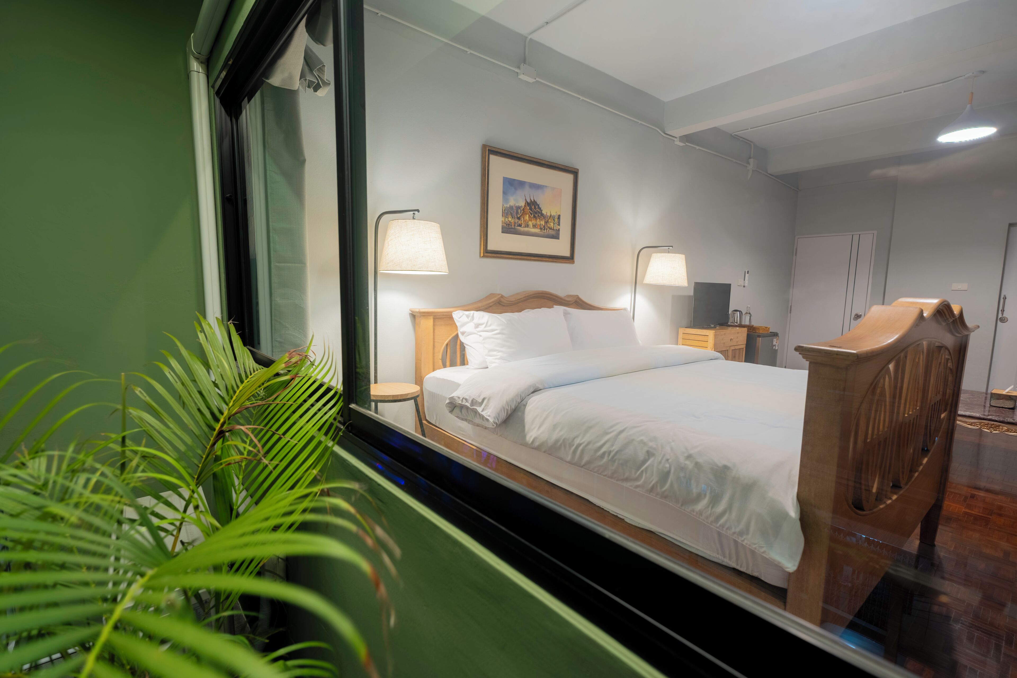 A hotel room with a bed, bedside lamps, a nightstand, a wooden headboard, a framed picture on the wall, and a green plant.