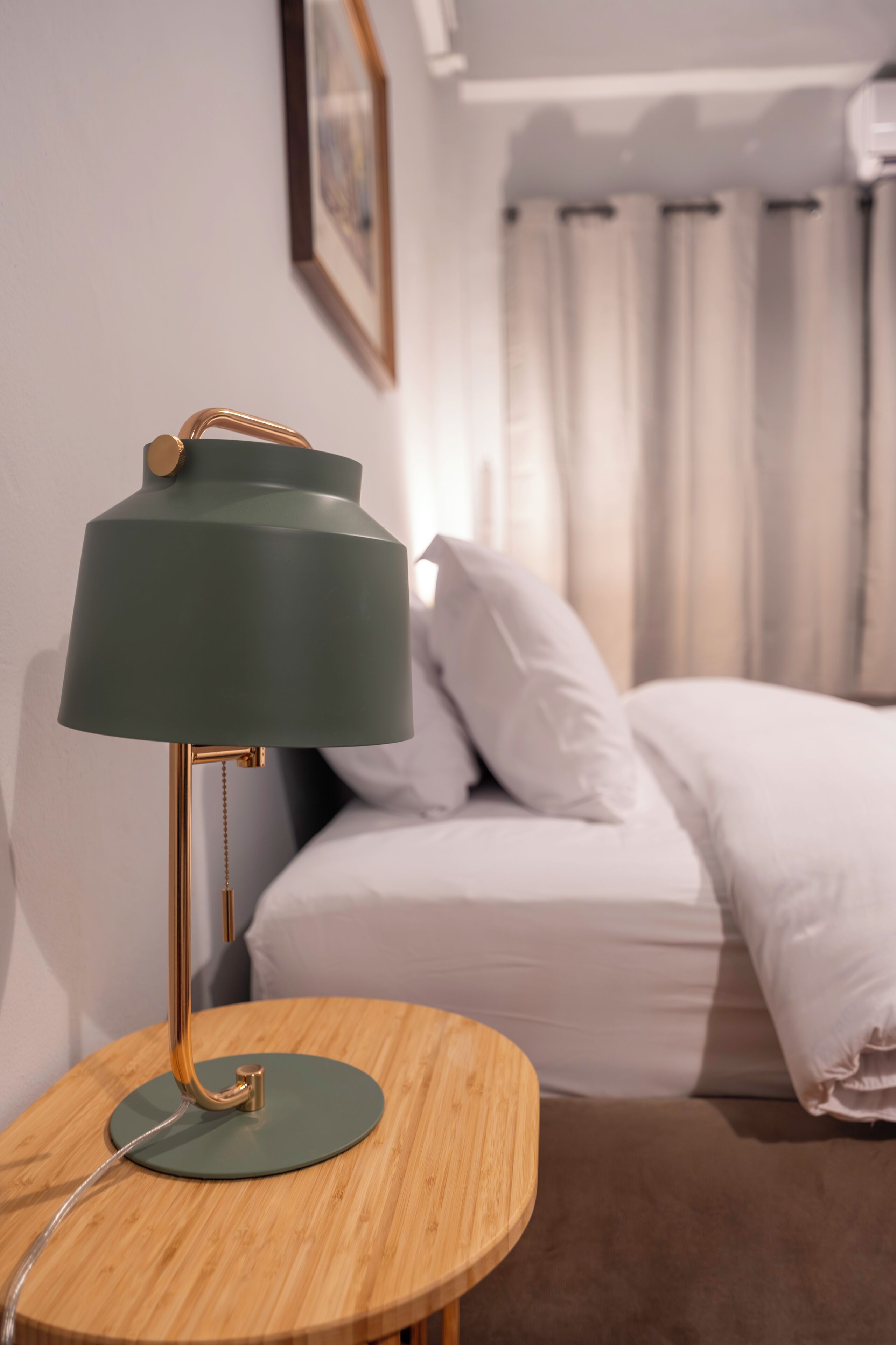 A round wooden table with a green lamp, a bed with white bedding, and a framed picture on the wall.