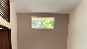 A corner room with a beige wall, a window with a view of foliage, a grey sofa, and a wooden door.