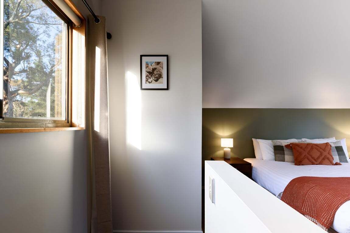 Une chambre à coucher avec un lit, une table de chevet avec une lampe, un tableau encadré au mur et une fenêtre avec des rideaux.