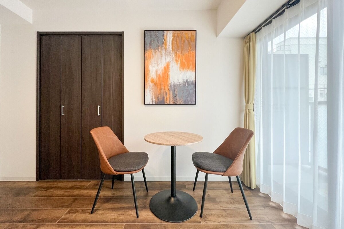 A room with a dark wooden wardrobe, two cushioned chairs, a round wooden table, and a large abstract painting on the wall.