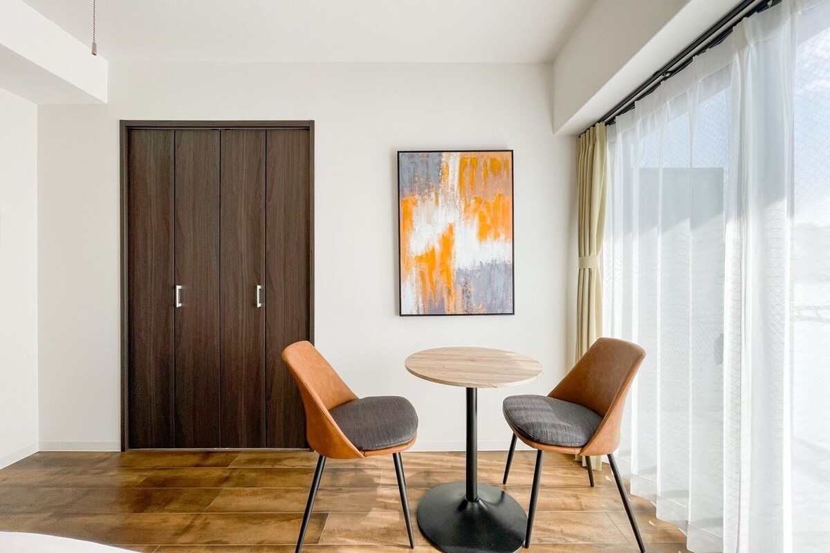 A room with a wooden wardrobe, two chairs, a round table, and a large abstract painting.