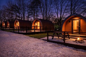 A row of wooden pod-style cabins with a gravel path in between, each with a bench and a fire pit. - Wyreside pod, ideal family break on fishing park close to Lancaster and Blackpool (Dolphinholme)