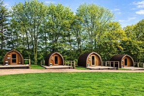 Tres cabañas de madera con techos abovedados, cada una con una puerta y un porche pequeño, rodeadas de césped y árboles.