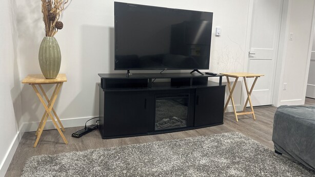 Uma sala de estar com uma televisão de tela plana em um suporte preto, uma mesa lateral de madeira, um tapete cinza e uma porta ao fundo.