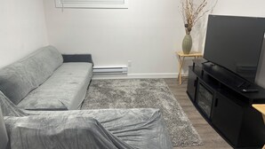 Uma sala de estar com um sofá cinza, uma TV de tela plana sobre um suporte preto, uma pequena mesa lateral de madeira e um tapete cinza.