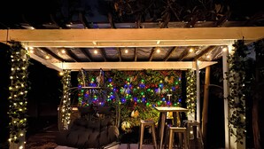 A covered outdoor seating area with a cushioned chair, a table, and chairs, surrounded by greenery and string lights. - Mid-Term Luxury Family Stay/Hot Tub, 2 Fire Pits, Sauna, Massage Chair & Arcade (Alexandria)