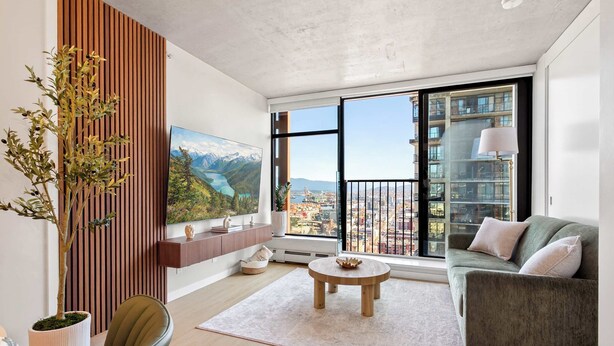 Un salon moderne comprenant un canapé, une table basse ronde, une plante en pot, un téléviseur fixé au mur et une grande fenêtre donnant sur la ville.