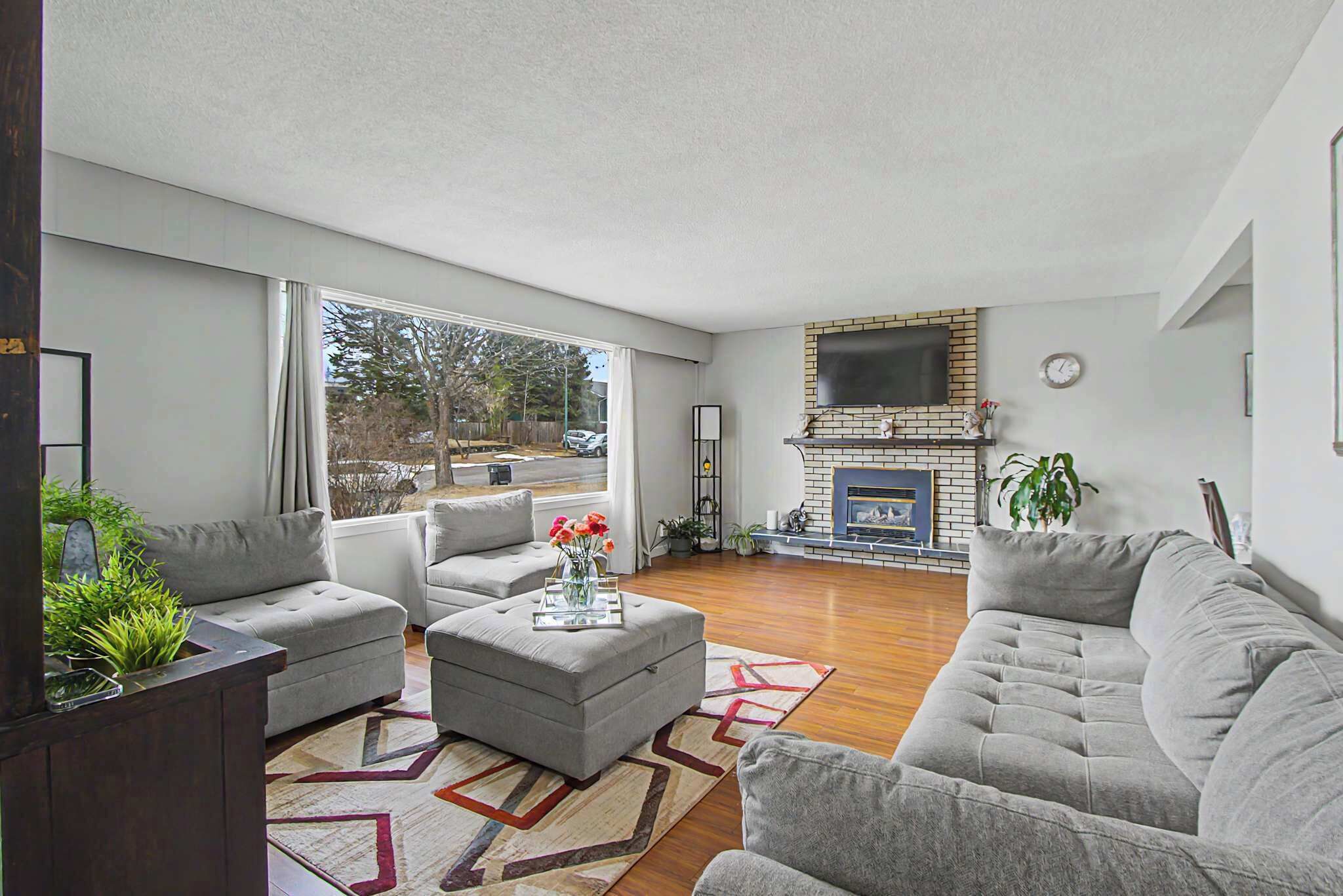 Bright, open-concept living room with rich hardwood floors and a large picture window. Features a cozy grey sectional, a geometric area rug, and a classic brick fireplace with a mounted flat-screen TV.