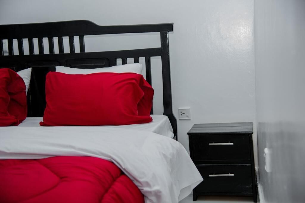A bed with a black headboard, red pillows, and a white bedspread. There is a small black nightstand with drawers to the right of the bed.
