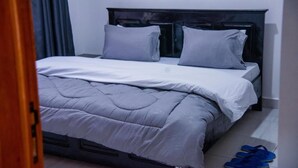 Un lit bien fait, avec une couette et des oreillers gris, une tête de lit en bois et une paire de pantoufles bleues posées sur le sol.