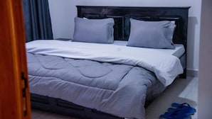 A neatly made bed with a grey duvet and pillows, a wooden headboard, and a pair of blue slippers on the floor. - Vista Homes Villa (Kigali)