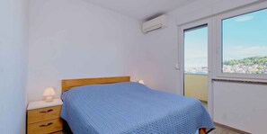 A bedroom with a bed, a nightstand, a lamp, a window with a view of a town, and an air conditioning unit. - Holiday Apartment Frantin A2 - A (Mali Lošinj)