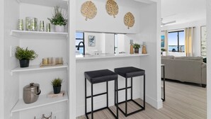 Une cuisine moderne avec des meubles blancs, un plan de travail noir et une vue sur la mer. On y trouve deux tabourets de bar noirs, une plante en pot et un élément décoratif en forme de feuille accroché au mur.