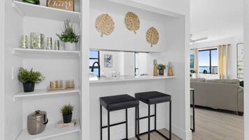 A modern kitchen with white cabinetry, a black countertop, and a view of the sea. There are two black bar stools, a potted plant, and a decorative leaf wall piece.