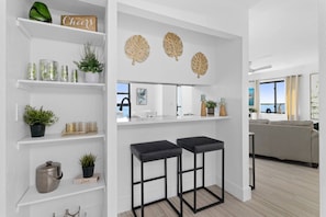 Una cocina moderna con armarios blancos, encimera negra y vista al mar. Hay dos taburetes negros, una planta en maceta y un elemento decorativo de hojas en la pared.