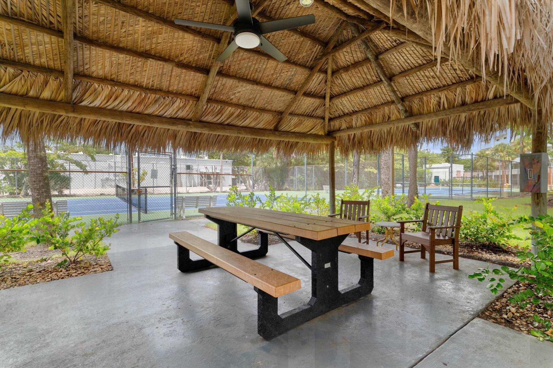 An outdoor pavilion with a thatched roof, a table, benches, and a view of a tennis court.
