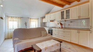 A cozy living room with a sofa and a small table, a kitchen with white cabinets and a tiled backsplash, and a window with curtains. - Holiday Apartment Biserka B4 B (Cunski)