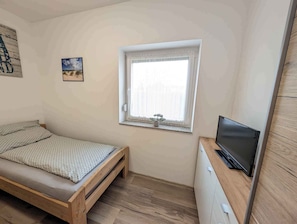 Un dormitorio pequeño con una cama individual, un televisor sobre un aparador de madera, una ventana con vistas y una planta pequeña en el alféizar.