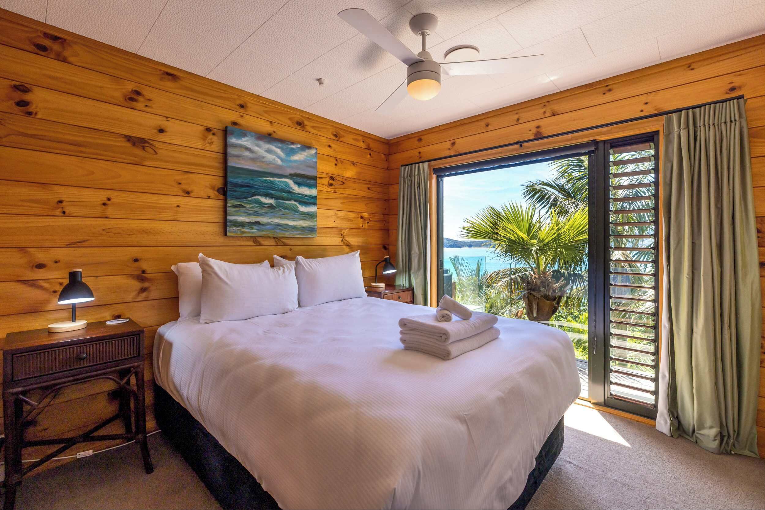A hotel room with a wooden interior, a bed with white bedding, a ceiling fan, a nightstand with a lamp, and a view of palm trees through a sliding glass door.
