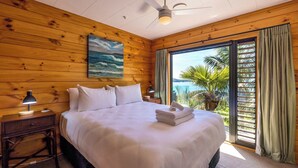 A hotel room with a wooden interior, a bed with white bedding, a ceiling fan, a nightstand with a lamp, and a view of palm trees through a sliding glass door. - Palms on Kennedy Point (Waiheke Island)