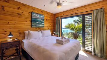 A hotel room with a wooden interior, a bed with white bedding, a ceiling fan, a nightstand with a lamp, and a view of palm trees through a sliding glass door.