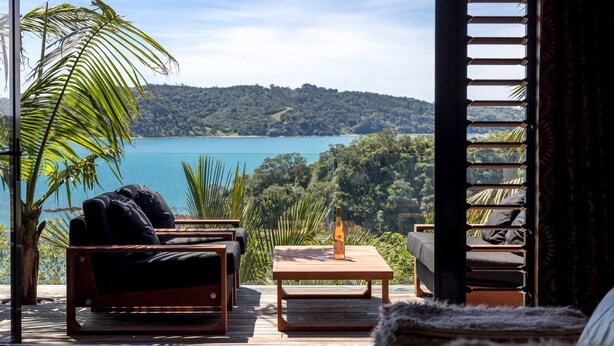 A wooden deck with outdoor furniture, a bottle of wine, and a view of a lake and forest. - Palms on Kennedy Point (Waiheke Island)