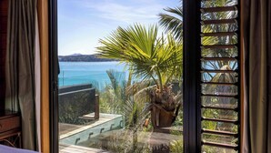 A room with a large window overlooking a body of water, a wooden deck, and a potted plant. - Palms on Kennedy Point (Waiheke Island)