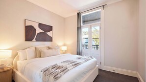 Quarto com cama grande, duas luminárias de cabeceira, um quadro na parede e vista através de uma porta.