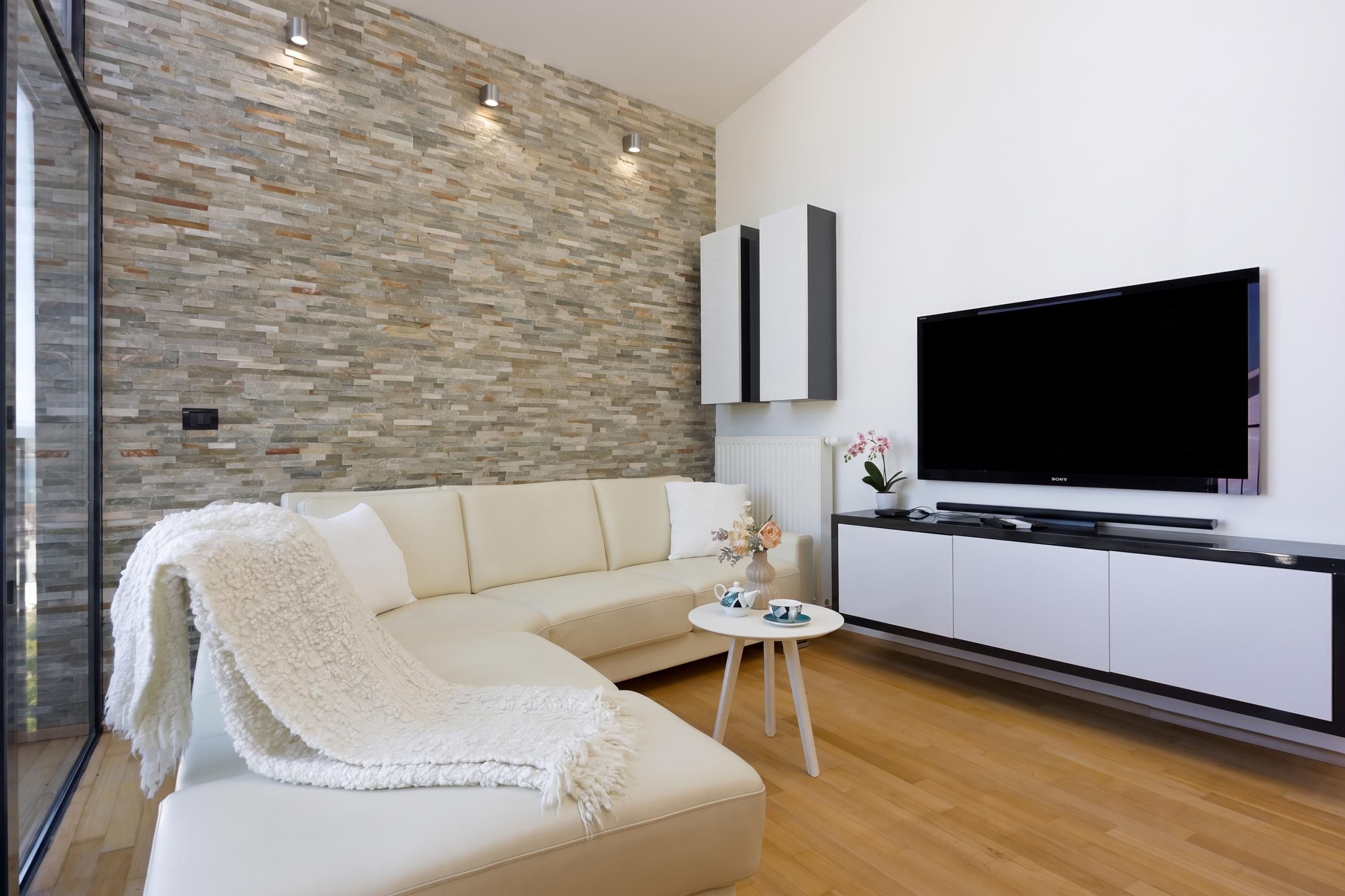 Ein modernes Wohnzimmer mit Steinwand, einem weißen Sofa, einem Flachbildfernseher und einem kleinen Beistelltisch.