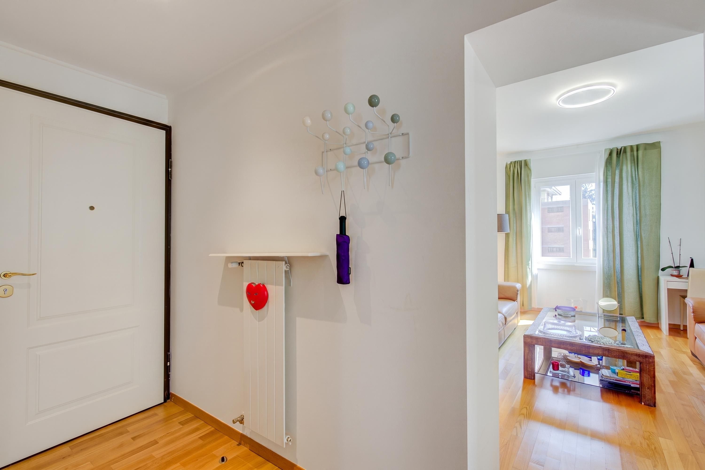 A room with a white door, a radiator, a heart-shaped decoration, a wall-mounted light fixture, and a glimpse of a living area with a sofa and a table.