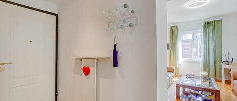 Una habitación con una puerta blanca, un radiador, una decoración con forma de corazón, una lámpara de pared y una vista parcial de una sala de estar con un sofá y una mesa.