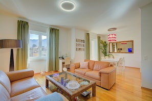 A living room with a sofa, a coffee table, a dining area with a table and chairs, a mirror, and a view of the outside. - Casa Berna - Balcony Apt With Mountain Views (Lugano)