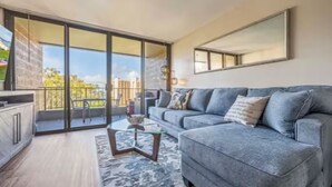 A modern living room with a grey sofa, a glass coffee table, and a large sliding glass door leading to a balcony.