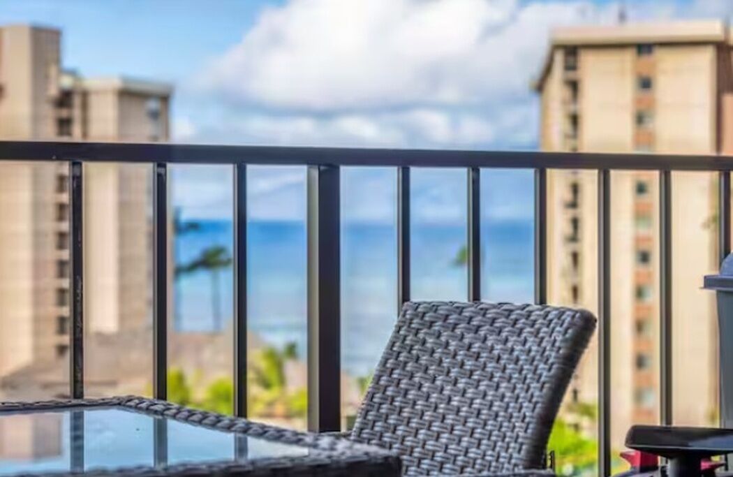 Un balcon avec un fauteuil en osier, une table en verre et une vue sur de hauts immeubles et l’océan.