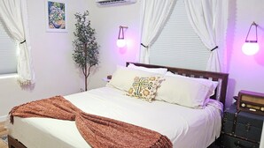 Un dormitorio con cama, cortinas, una lámpara de pared, una planta y una maleta.