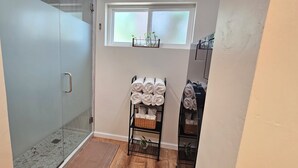 A bathroom with a glass shower enclosure, a towel rack with neatly rolled towels, a small shelf with toiletries, and a window above the shelf.