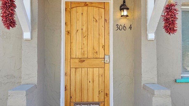 A building entrance with a wooden door, a number '306 4' on the wall, and a doormat with a wreath design.