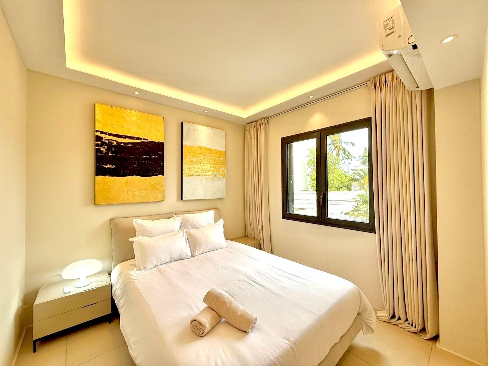 A hotel room with a bed, two paintings, a nightstand, a window with curtains, and an air conditioning unit.
