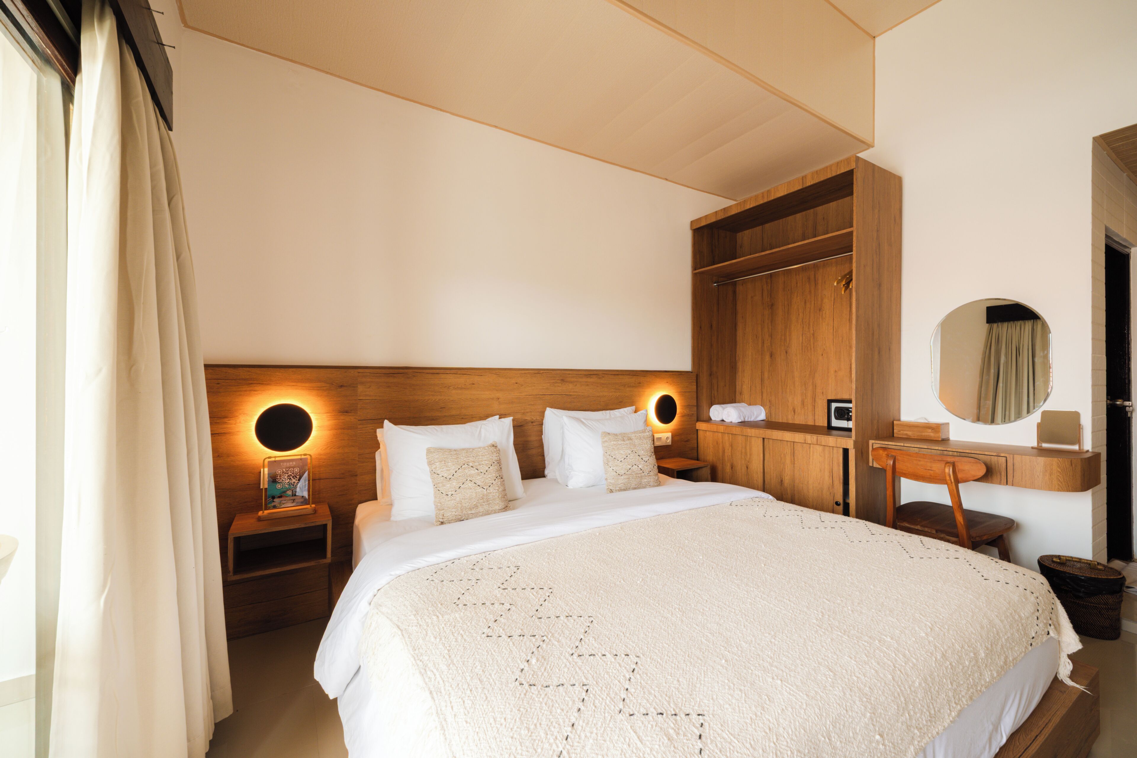 A bedroom with a large bed, wooden headboard, bedside tables with lamps, a wooden wardrobe, a mirror, and a small desk with a chair.