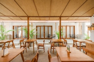 Un amplio comedor interior con mesas y sillas de madera, ventanas grandes y plantas en macetas.