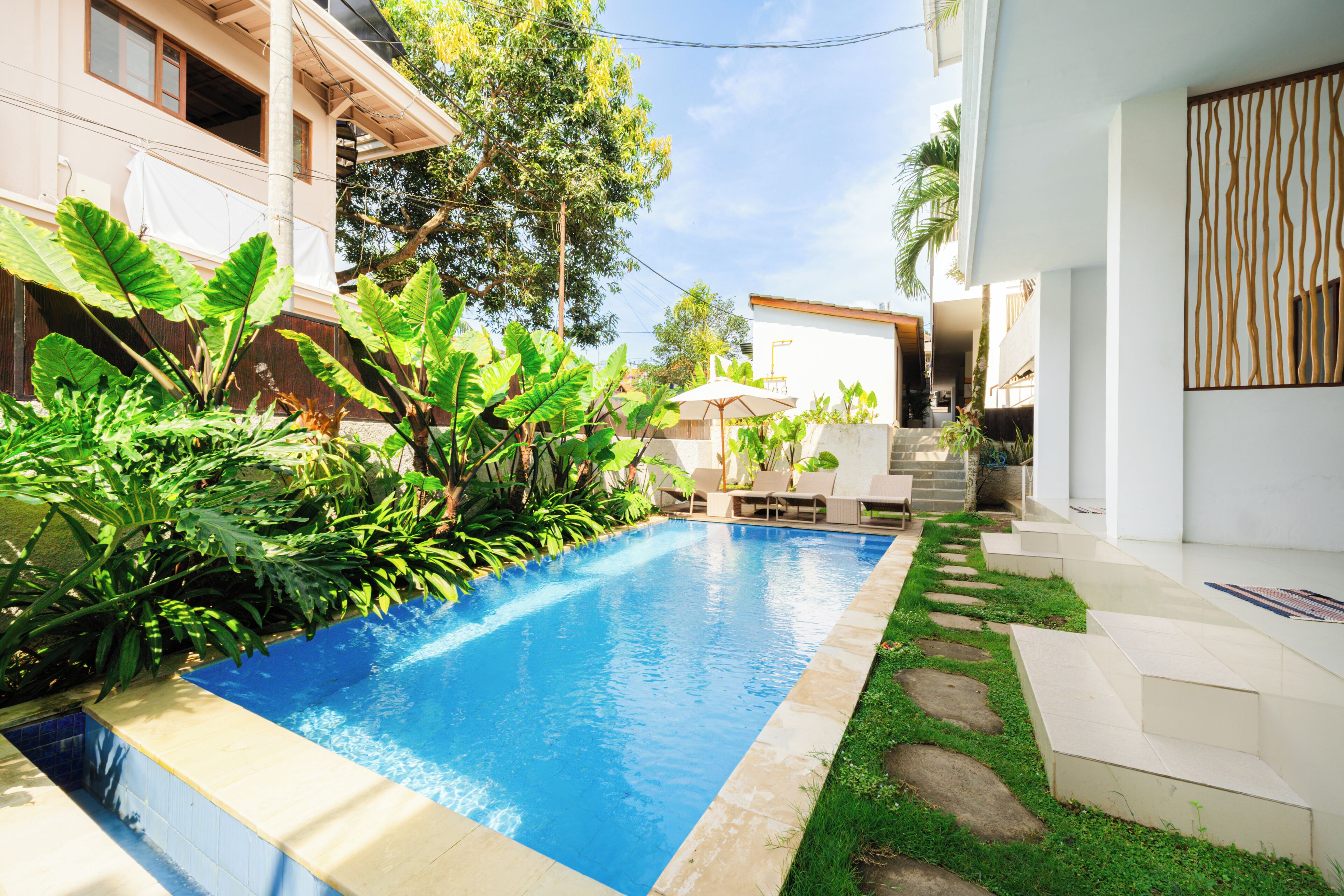 A swimming pool with steps leading to a covered area, surrounded by lush greenery and a white building.