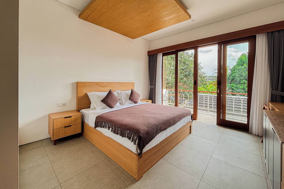 Quarto com cama de madeira, mesa de cabeceira, grande janela com vista para vegetação e luminária no teto.