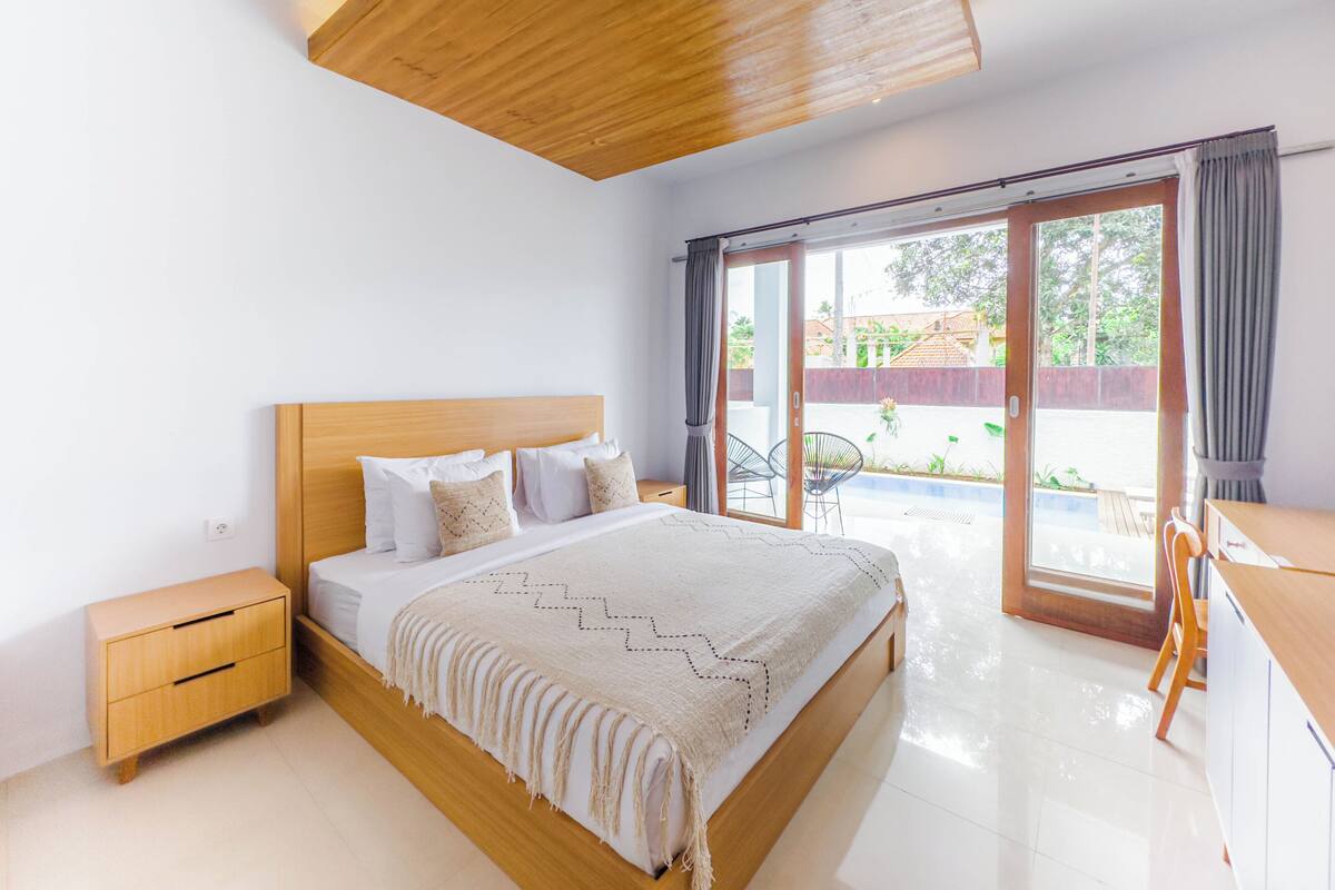 Quarto com cama de madeira, mesa de cabeceira, escrivaninha e porta de vidro que se abre para área externa.